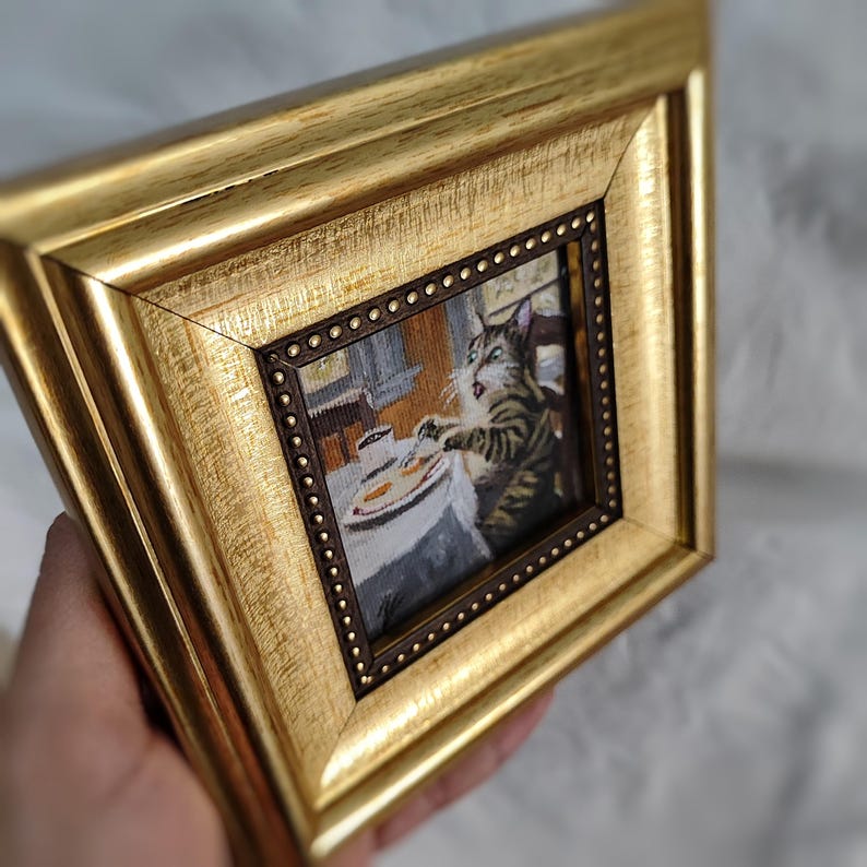 May include: A framed needlepoint artwork depicting a cat sitting at a table with a plate of food and a cup of coffee. The cat is looking at the food with a surprised expression.