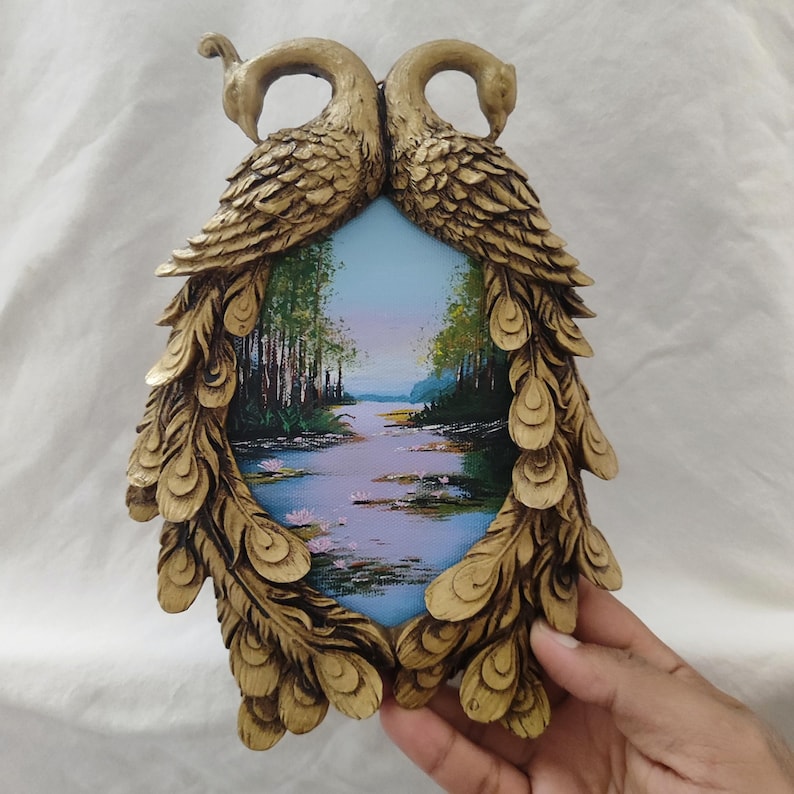 May include: A decorative wall art piece featuring a painting of a river scene framed by two golden peacock figures. The frame is intricately detailed with feather-like patterns. The painting depicts a serene landscape with trees and water.