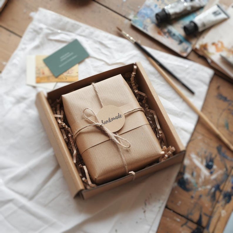 May include: A brown cardboard box containing a wrapped gift. The gift is wrapped in brown paper and tied with twine, with a circular tag that reads "handmade". The box sits on a white cloth, with art supplies in the background.