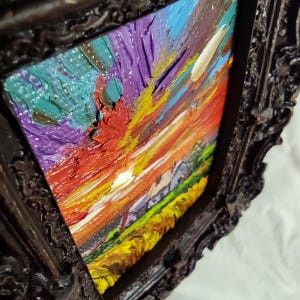 May include: An abstract oil painting in a dark brown ornate frame. The artwork features a vibrant sunset with bold brushstrokes of red, orange, yellow, blue, and purple. The textured paint creates a three-dimensional effect.
