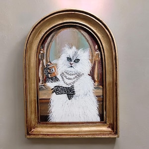 May include: A painting in a gold-coloured frame, depicting a fluffy white cat wearing a pearl necklace and black gloves, holding a small object. The cat is in front of a mirror. The painting has a vintage aesthetic and is in an arched frame.