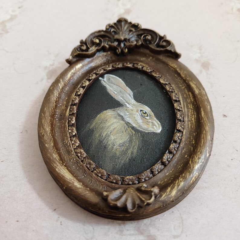 May include: An oval-shaped, antique-style frame with an elaborate design. Inside the frame is a small painting of a rabbit with a light brown and white coat, set against a dark background. The frame has a bronze finish.