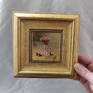 Peut inclure: Petite peinture encadr&eacute;e carr&eacute;e d'un cocktail dans un verre &agrave; martini avec une cerise et des olives. Le cadre est dor&eacute; et la peinture a un fond dor&eacute;. La peinture est tenue par une main.