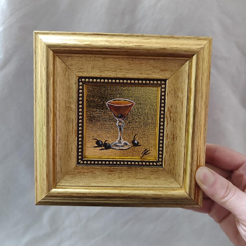 Peut inclure: Petite peinture encadr&eacute;e carr&eacute;e d'un cocktail dans un verre &agrave; martini avec une cerise et des olives. Le cadre est dor&eacute; et la peinture a un fond dor&eacute;. La peinture est tenue par une main.