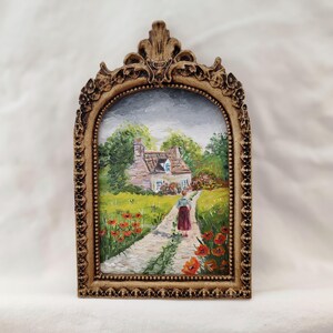 May include: A framed miniature painting of a cottage scene. The artwork depicts a person walking on a path towards a white cottage with a red roof, surrounded by green trees and red poppies. The frame is ornate, with a gold-colored finish.