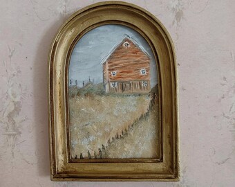 May include: An arched gold-toned frame containing a painting of a rustic orange barn situated in a dry, golden-brown field. The scene includes a small wooden fence line leading toward the structure under a pale, overcast sky with muted gray tones.