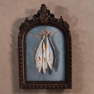 Op de afbeelding: Een schilderij in een sierlijke lijst toont drie vissen die aan een touwtje hangen tegen een blauwe achtergrond. De vissen zijn geschilderd in grijstinten, bruin en wit. De lijst is donkerbruin.