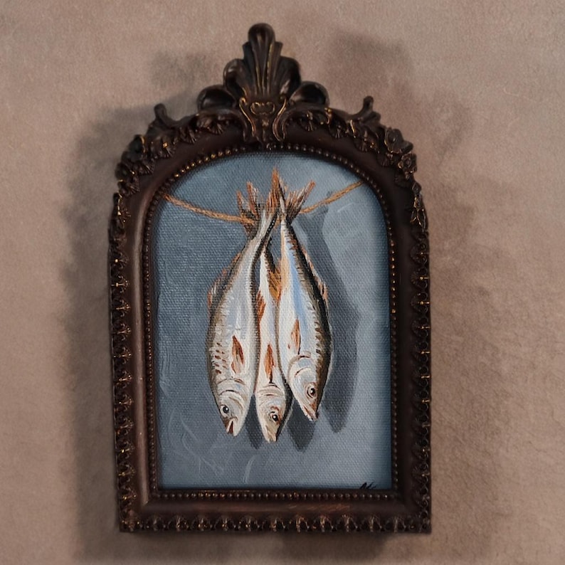 Op de afbeelding: Een schilderij in een sierlijke lijst toont drie vissen die aan een touwtje hangen tegen een blauwe achtergrond. De vissen zijn geschilderd in grijstinten, bruin en wit. De lijst is donkerbruin.