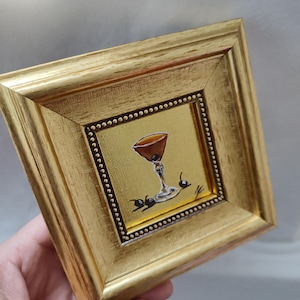 Peut inclure: Petite peinture encadr&eacute;e carr&eacute;e d'un cocktail dans un cadre dor&eacute;. La peinture repr&eacute;sente un cocktail brun dans un verre &agrave; pied avec deux cerises. Le cadre est dor&eacute; avec une finition textur&eacute;e et une bordure int&eacute;rieure pointill&eacute;e.