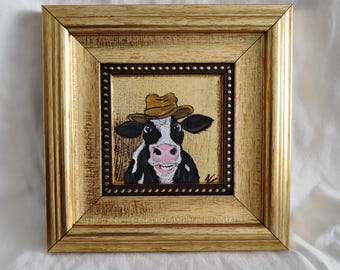 May include: A framed painting of a black and white cow wearing a brown hat. The cow is smiling and has a pink nose. The painting is set against a gold background.