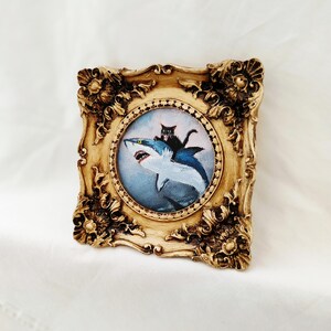 May include: Ornate, gold-colored framed artwork featuring a whimsical painting of a black cat riding a blue and white shark. The artwork has a circular painting with a detailed, decorative frame.