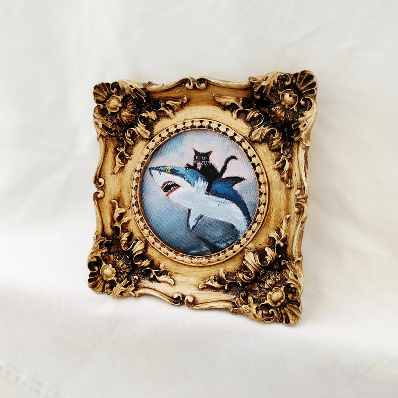 May include: Ornate, gold-colored framed artwork featuring a whimsical painting of a black cat riding a blue and white shark. The artwork has a circular painting with a detailed, decorative frame.