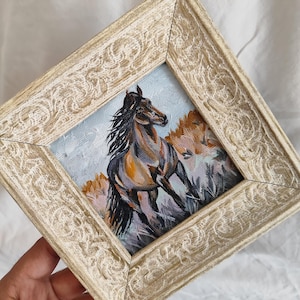 May include: A framed painting depicting a running horse. The artwork shows a brown and grey horse with a flowing mane, set against a light blue sky and brown hills. The frame is a distressed cream colour.