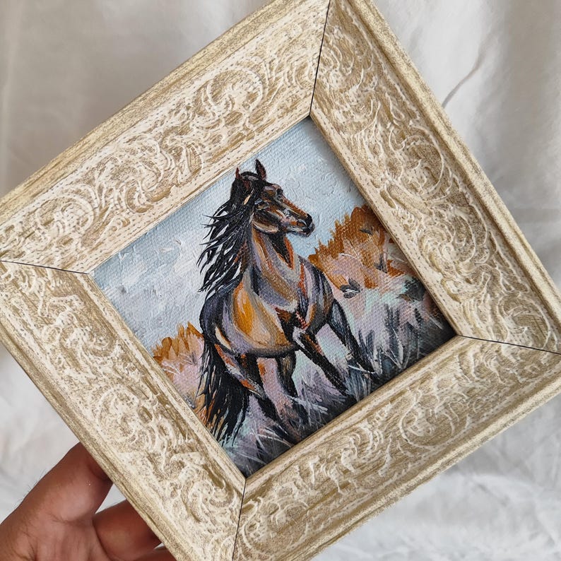 May include: A framed painting depicting a running horse. The artwork shows a brown and grey horse with a flowing mane, set against a light blue sky and brown hills. The frame is a distressed cream colour.