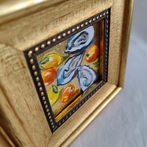 K&ouml;nnte beinhalten: Ein kleines, gerahmtes Gem&auml;lde mit einem goldfarbenen Holzrahmen. Das Kunstwerk zeigt Austern und Zitrusfr&uuml;chte in Blau-, Orange- und Gelbt&ouml;nen. Der Rahmen hat einen dunkelbraunen Innenrand mit kleinen, goldfarbenen Nieten.