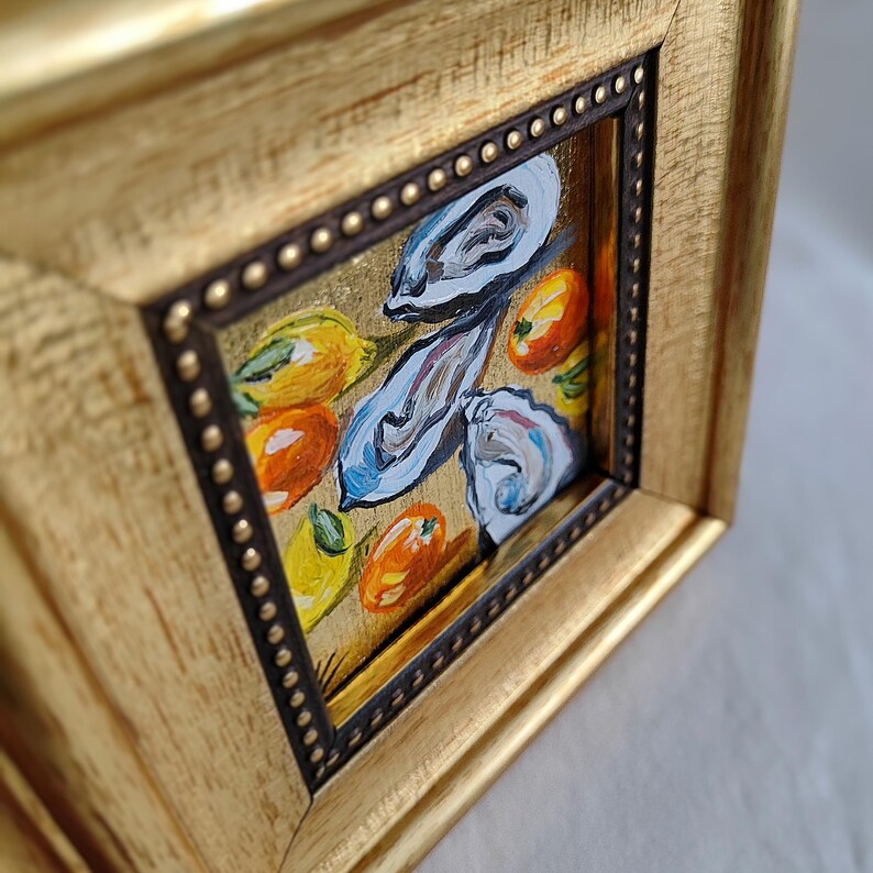 K&ouml;nnte beinhalten: Ein kleines, gerahmtes Gem&auml;lde mit einem goldfarbenen Holzrahmen. Das Kunstwerk zeigt Austern und Zitrusfr&uuml;chte in Blau-, Orange- und Gelbt&ouml;nen. Der Rahmen hat einen dunkelbraunen Innenrand mit kleinen, goldfarbenen Nieten.