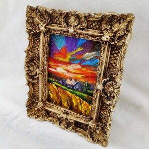 May include: A small framed painting depicting a sunset over a rural landscape. The artwork features a vibrant sky with orange, purple, and blue hues, and a field of yellow and green. The ornate frame is a light beige color.