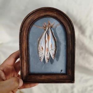 Op de afbeelding: Een ingelijst schilderij van drie vissen die aan een touwtje hangen. De vissen zijn zilverkleurig met blauwe en witte accenten, tegen een lichtblauwe achtergrond. De lijst is donkerbruin en boogvormig. Het kunstwerk is een stilleven.