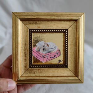 Könnte beinhalten: Ein kleines gerahmtes Gemälde eines weißen Hundes, der auf einem rosafarbenen Koffer schläft. Das Gemälde ist auf goldenem Hintergrund.
