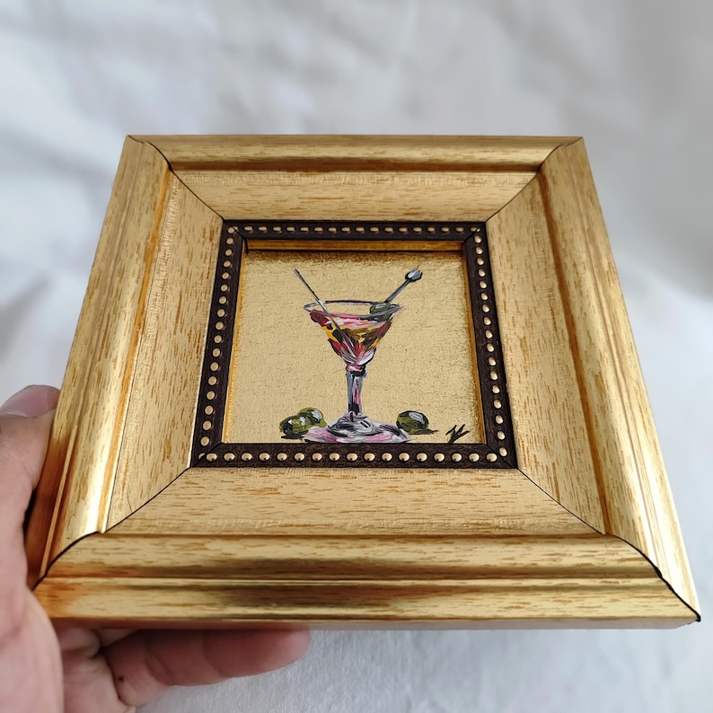 May include: A small framed painting of a martini glass with olives and a gold background.