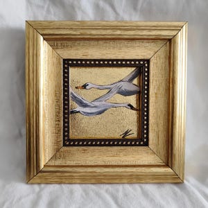 May include: A framed painting of two white swans in flight against a gold background. The painting is in a gold frame with a dark brown inner border.