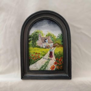 May include: A framed painting of a cottage scene. A person in a red skirt walks down a path toward a cottage with a white facade and blue shutters. Red poppies line the path. The painting is in a dark arched frame.