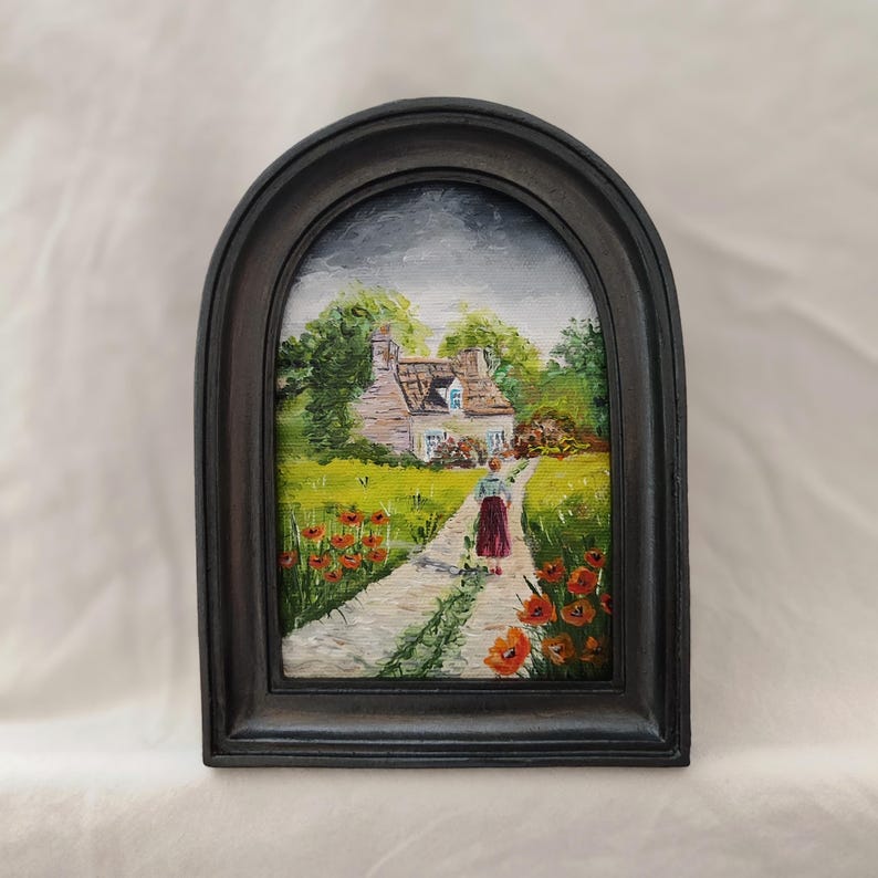 May include: A framed painting of a cottage scene. A person in a red skirt walks down a path toward a cottage with a white facade and blue shutters. Red poppies line the path. The painting is in a dark arched frame.