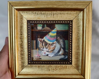 May include: A framed painting of a cat wearing a colorful party hat, looking sadly at a small cupcake with a lit candle. The cat is brown, white, and black. The frame is gold with a brown inner border.