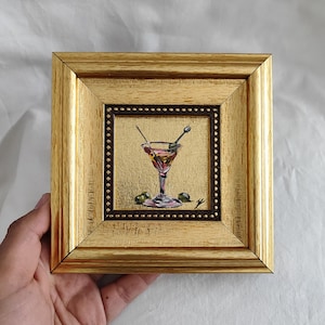 May include: A small framed painting of a martini glass with olives and a stirrer. The painting is in a gold frame with a brown mat.