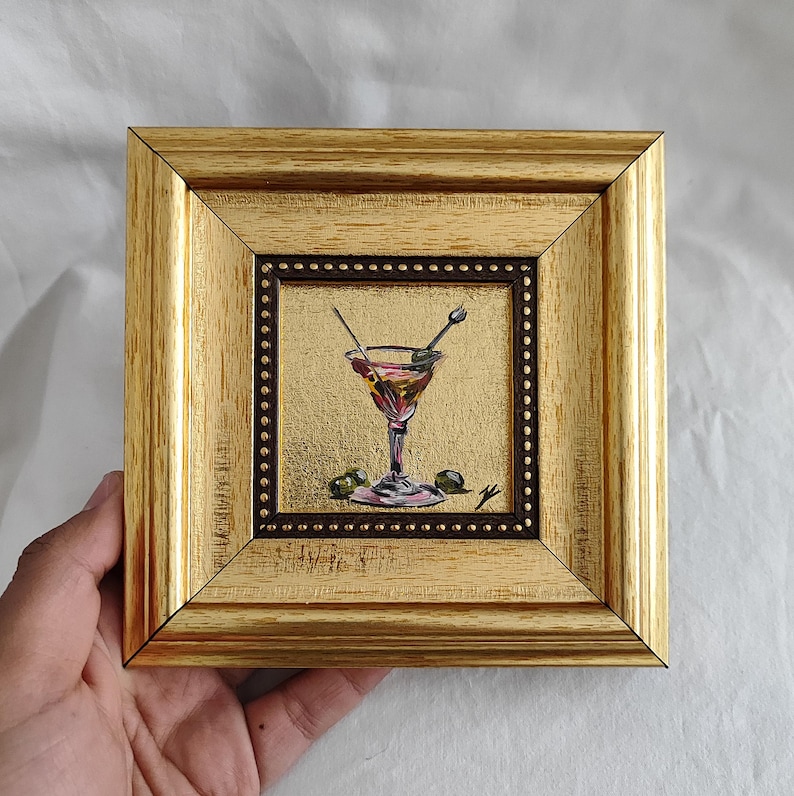 May include: A small framed painting of a martini glass with olives and a stirrer. The painting is in a gold frame with a brown mat.