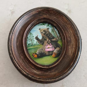 May include: A framed painting of a brown bear sitting in a forest setting. The bear is wearing a pink purse and has a finger raised in the air. The painting is in an oval frame.