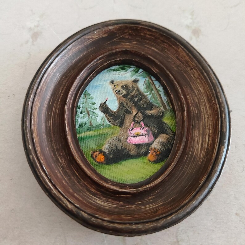 May include: A framed painting of a brown bear sitting in a forest setting. The bear is wearing a pink purse and has a finger raised in the air. The painting is in an oval frame.