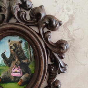 May include: A framed painting of a brown bear wearing lipstick and holding a pink purse. The bear is sitting in a green grassy area with trees in the background.