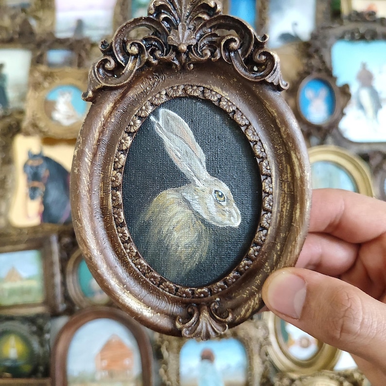 May include: An oval painting of a hare in a bronze-coloured, ornate frame. The hare is painted with detailed fur and a focused gaze against a dark background. The frame has an elaborate, vintage design. The painting is surrounded by other framed artworks.