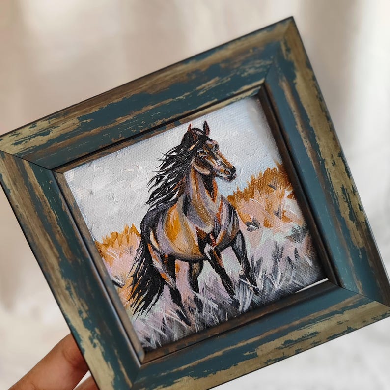 May include: A framed painting of a running horse. The horse has a brown and black coat, set against a backdrop of tall grass and a light sky. The frame is a distressed teal colour.