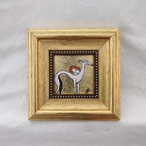May include: A framed artwork featuring a stylized illustration of a greyhound with a woman's head and red hair. The artwork is set against a gold background and framed in a gold-colored wooden frame. The frame has a dark brown inner border with small, evenly spaced dots.