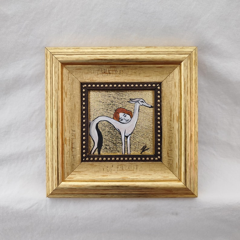 May include: A framed artwork featuring a stylized illustration of a greyhound with a woman's head and red hair. The artwork is set against a gold background and framed in a gold-colored wooden frame. The frame has a dark brown inner border with small, evenly spaced dots.