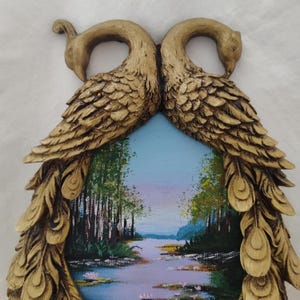 May include: A decorative wall art piece featuring a landscape painting framed by two gold-colored peacock figures. The painting depicts a river scene with trees and a blue sky. The peacock figures have detailed feather textures.