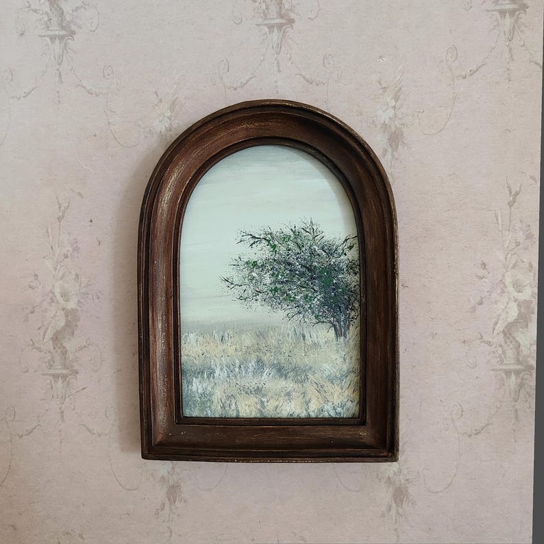 May include: A framed landscape painting featuring a tree with green foliage and a field of tall grass. The painting is set against a light sky and is enclosed in a dark brown, arched wooden frame. The artwork is mounted on a pink floral wallpaper.