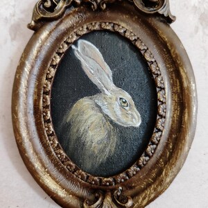 May include: An oval-shaped, antique-style frame featuring a painted portrait of a hare. The hare is rendered in brown and white tones against a dark backdrop. The frame is a distressed gold colour with intricate detailing, giving it a vintage feel.
