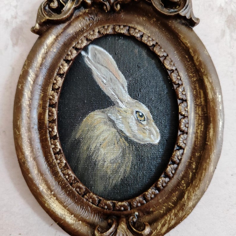 May include: An oval-shaped, antique-style frame featuring a painted portrait of a hare. The hare is rendered in brown and white tones against a dark backdrop. The frame is a distressed gold colour with intricate detailing, giving it a vintage feel.