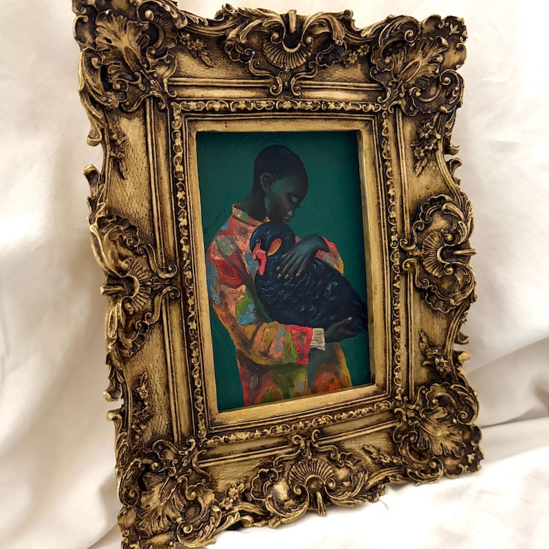May include: A rectangular painting in an ornate, antique-style gold frame. The artwork depicts a person with dark skin wearing a colorful, patterned shirt, holding a large black bird with a red beak against a solid dark teal background.