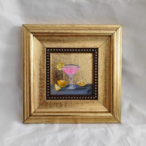 Peut inclure: Une petite peinture encadrée d'un cocktail rose dans un verre à martini avec des tranches de citron sur un fond doré.