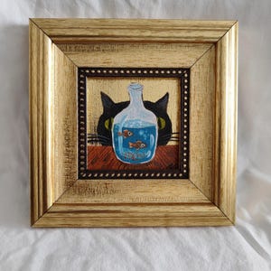 May include: A framed painting of a black cat peering over a glass bottle filled with blue water and three small orange and white fish. The cat has green eyes and a black nose.