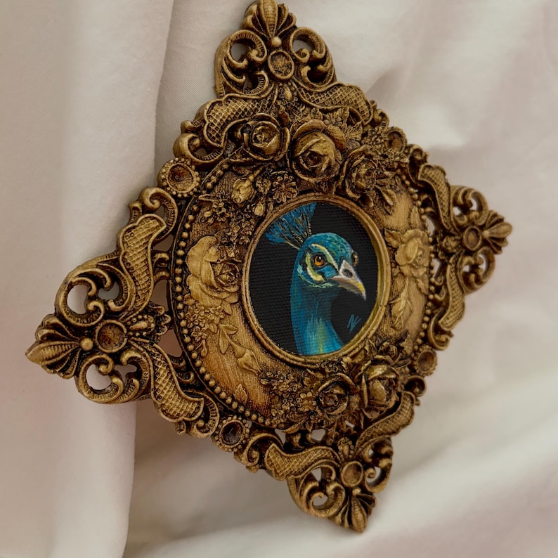 May include: A diamond-shaped wall hanging featuring a central circular painting of a peacock head with blue and green feathers. The artwork is encased in an ornate, metallic gold-colored frame decorated with sculpted floral and scrollwork details.
