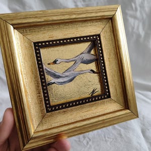 May include: A framed painting of two white swans in flight against a gold background. The swans are painted in a realistic style with detailed feathers. The frame is gold with a dark brown border.