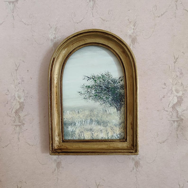 May include: A framed landscape painting featuring a tree with green foliage in a field of tall grass. The painting is set against a pale sky and is enclosed in a gold-colored, arched frame.