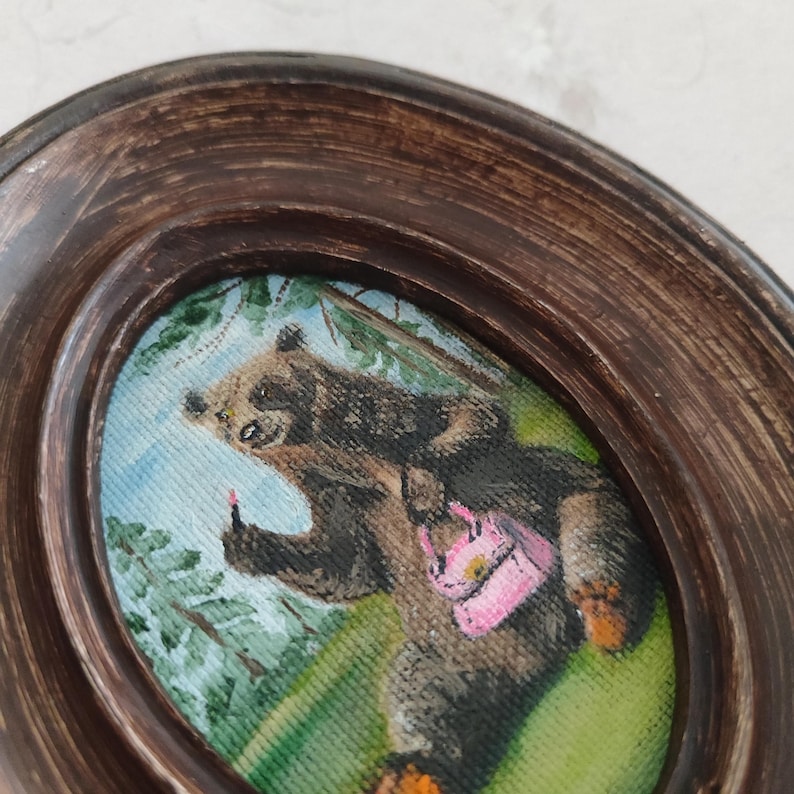 May include: A framed painting of a brown bear wearing a pink purse and giving the middle finger. The bear is standing in a green forest setting.