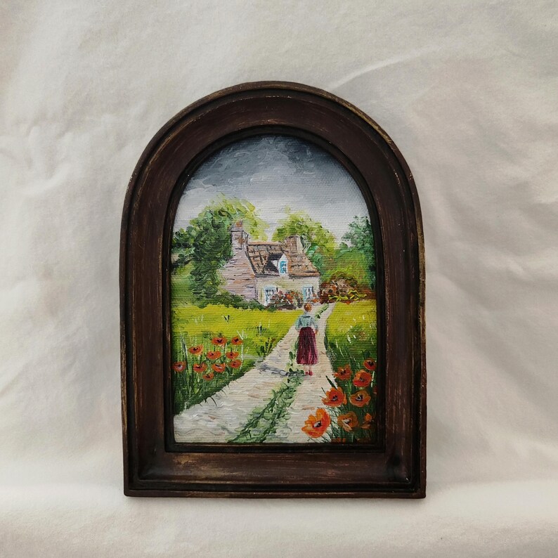 May include: A framed painting of a cottage scene. The artwork depicts a path leading to a house with a red roof, surrounded by green grass and red flowers. The frame is a dark brown, arched shape.