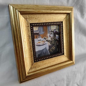 May include: A framed needlepoint artwork depicting a cat sitting at a table with a plate of food. The cat is looking at the food with a surprised expression. The frame is gold with a dark brown inner border.